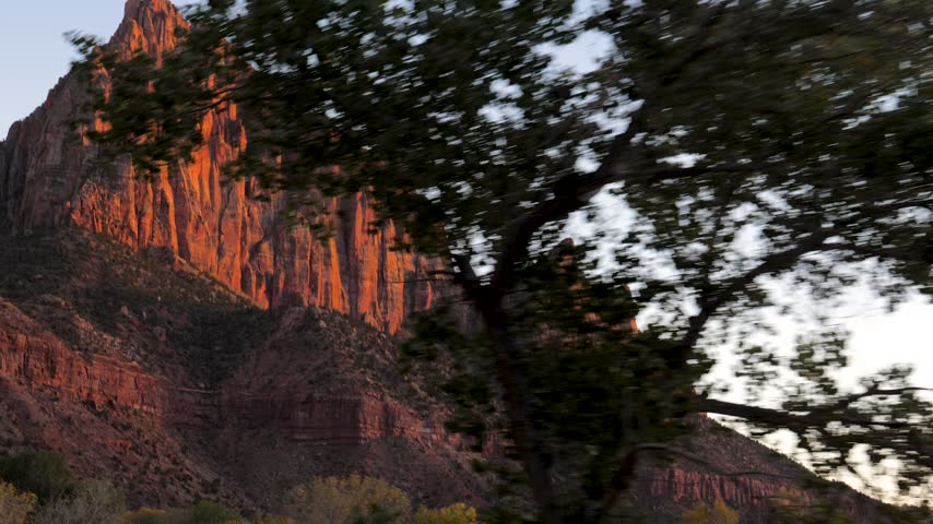 Scenic view from a moving car of the majestic red rock mountains in zion national park, utah, beautifully illuminated by the warm light of the setting sun against a clear blue sky