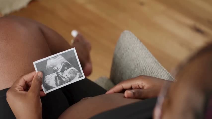 Pregnant African American woman sitting on a couch and lovingly looking at an ultrasound scan of her unborn child during the day at home in a warm and bright living space filled with anticipation