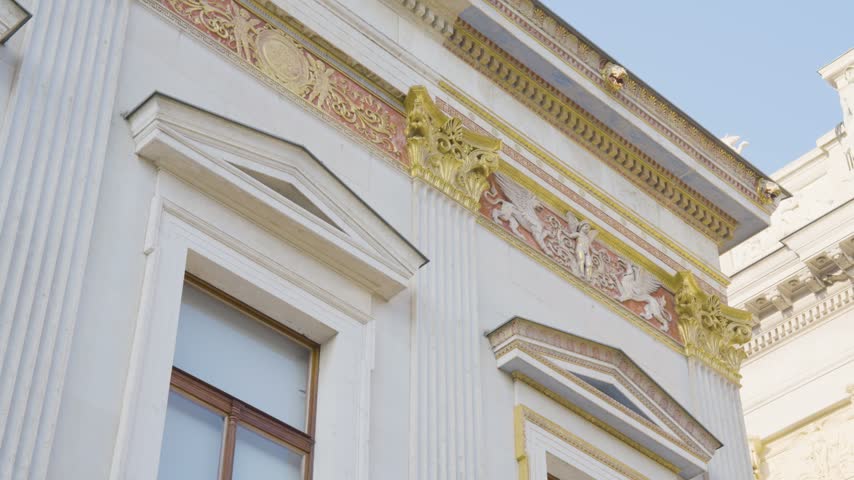 Close-up shot of the intricate golden and red frieze adorning the side facade of the Austrian Parliament Building in Vienna, featuring detailed relief carvings with mythological griffin and figure motifs above the windows, set against a clear blue summer sky.