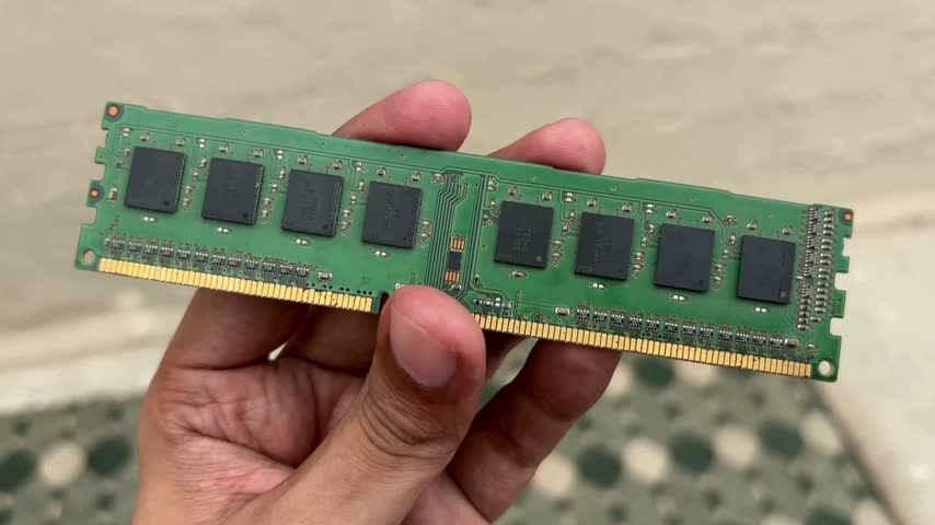 Close-up of a hand presenting a computer memory module or RAM. The module has multiple black chips on a green circuit board. Detail shot showing technology and hardware components.