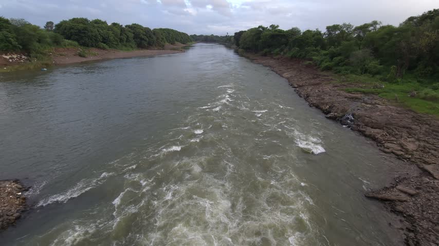 Wide angle panoramic view of a swelling river with white foam rapids extending towards the horizon under a cloudy sky, conceptualizing environmental changes, flood warning, and seasonal monsoon overflow
