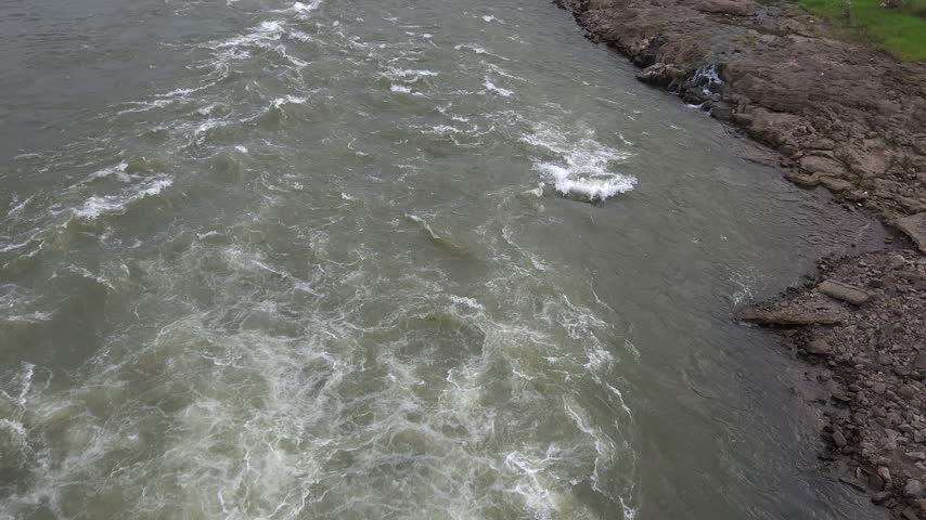 High angle view of turbulent white foam rapids rushing downstream along a rocky riverbed shore, capturing the immense power of nature and freshwater flow after heavy monsoon rains.