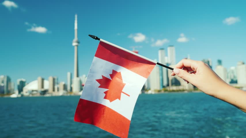 Close up view of hand holding and waving small Canadian flag against backdrop of downtown Toronto cityscape across water on bright sunny day with blue sky cinematic travel footage.