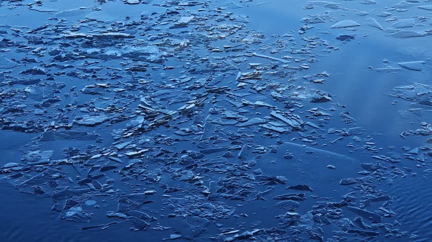 Close-up of spring thaw on reservoir. Small pieces of broken ice on smooth surface of blue water. River current carries thin layer of melting ice, winter, early spring. Natural background with ice