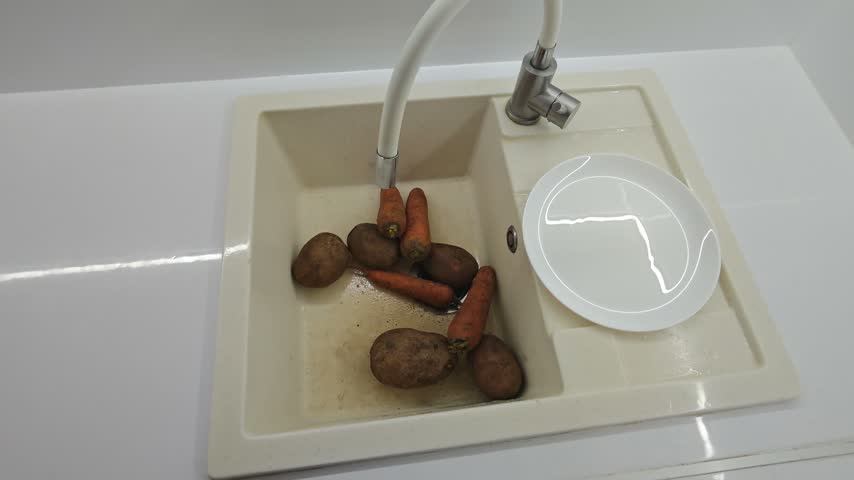 A top-down point of view shot of a person washing dirty fresh potatoes and carrots under running water in a modern beige kitchen sink before preparing a healthy meal.