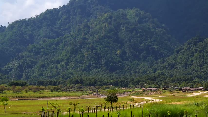 A scenic landscape featuring a densely forested mountain overlooking a wide green valley with rural fields, dirt paths, a small bridge, and countryside structures.