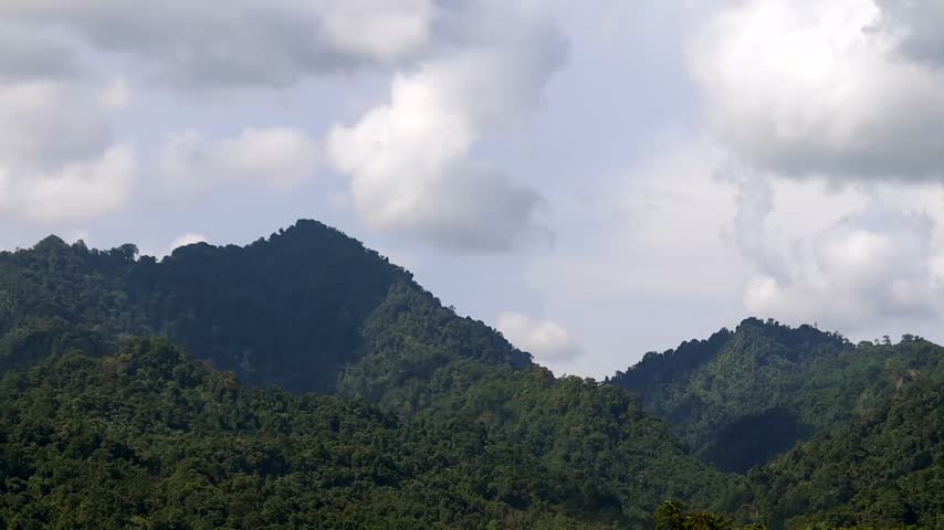 A scenic natural landscape featuring densely forested mountains with layered green hills beneath a bright sky filled with soft white and gray clouds.