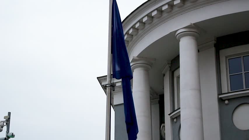 The blue European Union flag flutters on a pole in front of a historic white building featuring large columns and windows