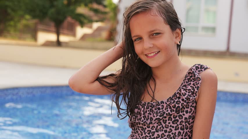 Portrait of a smiling young girl in a leopard swimsuit touching her wet hair while standing by a swimming pool on vacation.