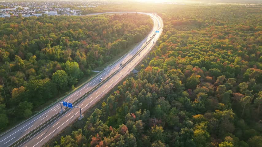 Curved divided highway through autumn forest in golden light with moving vehicles, overhead directional signs, and central barrier near suburban edge at sunset