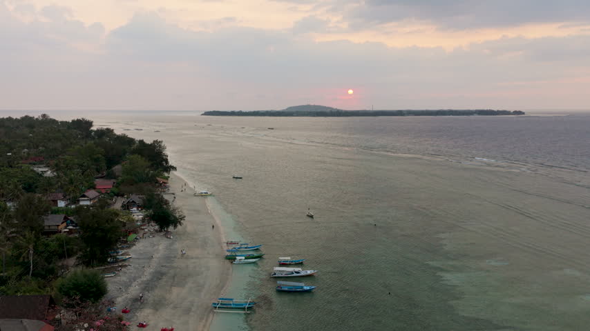 Aerial drone shot pulling backward along the coastline of Gili Air, Indonesia during a soft sunset. Scenic high-angle view showing small boats anchored in shallow water, beach huts, and the sun setting directly over neighbouring Gili Trawangan island on the horizon.   