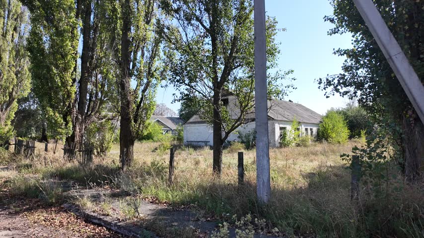 A dynamic tracking shot moving past an abandoned farmstead with a leaning wooden fence, overgrown grass, and old white rural houses under a bright sky.