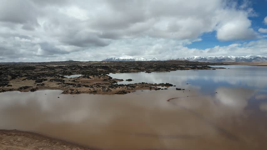 A wide view moves across shallow highland wetlands with exposed rocks, cloud reflections and distant snow-covered mountains.