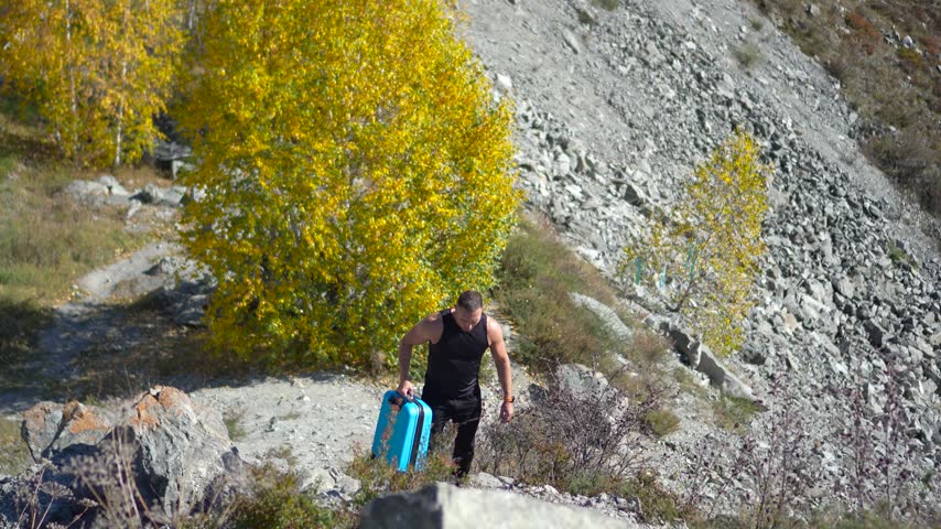 Athletic man carrying a blue suitcase up a steep rocky mountain path with a static camera shot.