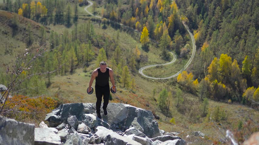 Athletic man climbing up rocky mountain pass holding weight plates with a static camera shot.