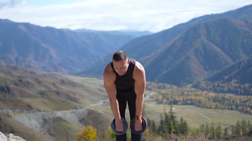 Athletic man holding weight plates out to the sides on a mountain pass with a static camera shot.
