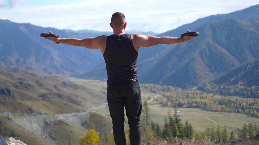 Athletic man holding weight plates out to the sides on a mountain pass with a static camera shot.
