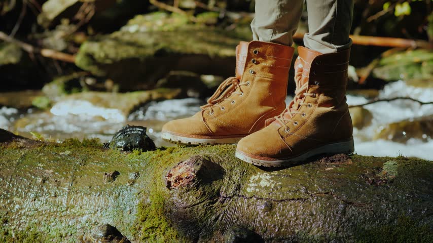 Close up of feet in durable leather hiking boots resting on a fallen tree trunk covered in green moss with a fast flowing creek in the background.