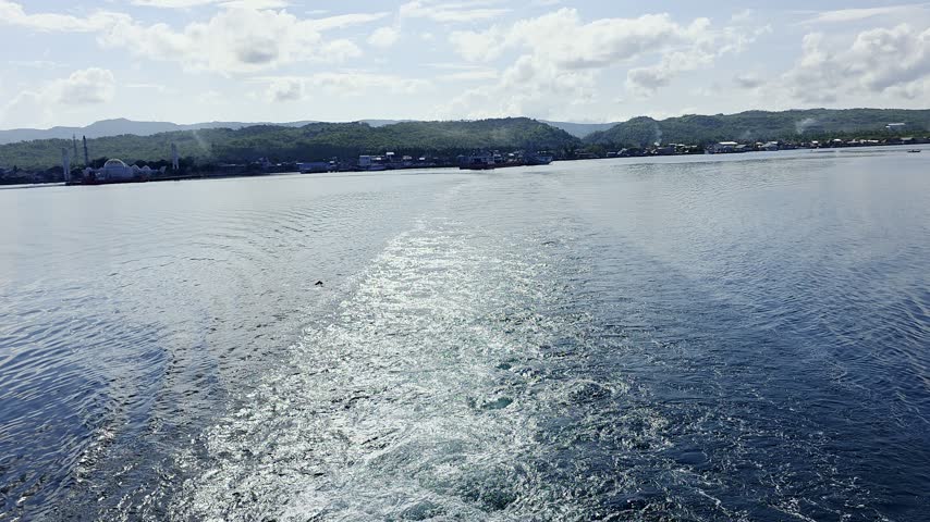 Ferry boat wake dynamic water waves trailing behind ship leaving coastal town under sunny sky.