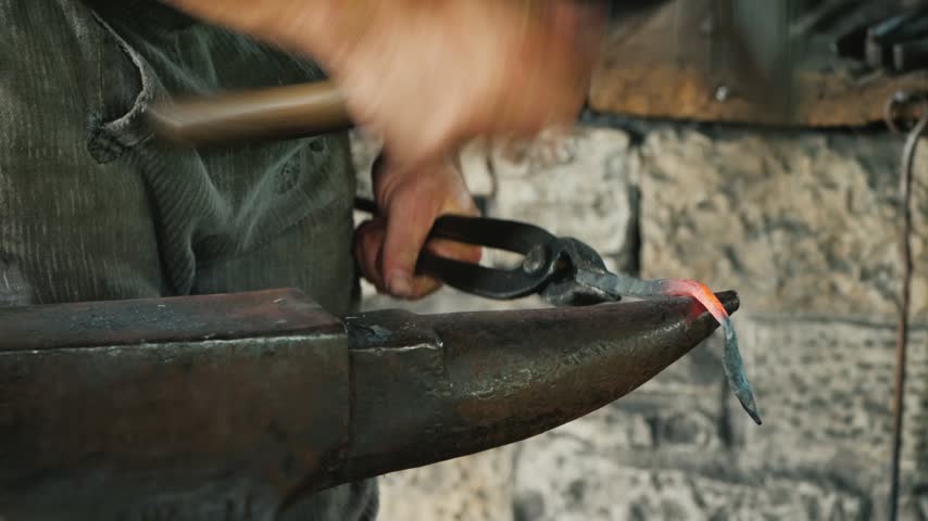 Skilled blacksmith working with glowing metal piece on iron anvil using hammer and tongs. Traditional craftsmanship scene showing metalworking process in rustic workshop environment.