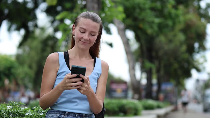 Smiling caucasian woman sitting on a bench in a city park, using her smartphone for texting, scrolling social media, or browsing the internet while traveling in Chiang Mai, Thailand