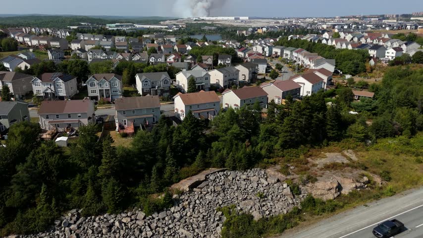A large wildfire spreads on the edge of Halifax, Nova Scotia. Thick smoke rises above the residential areas, casting a shadow over the landscape.