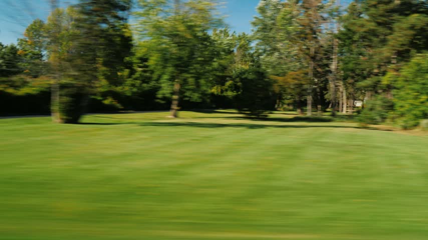 Dynamic motion blur captures rural scenery from moving vehicle window passing residential houses lush green lawns trees and clear blue sky during bright sunny autumn afternoon.