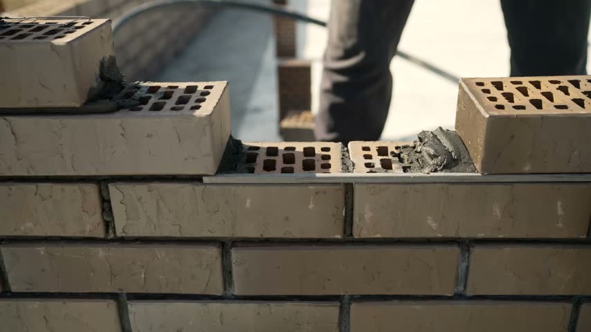 Adult man in white work gloves applying dark grey mortar with trowel to brown perforated brick while building new brick wall outdoors, construction work