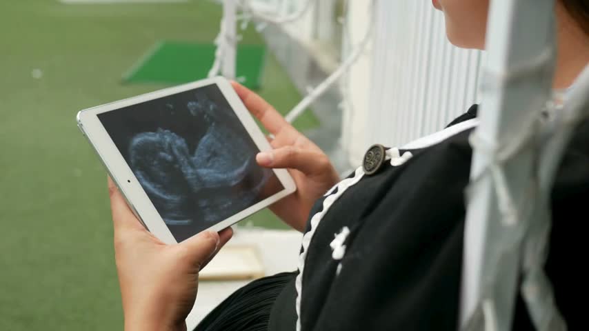 Expectant mother viewing her baby’s ultrasound on a tablet in a park garden for a prenatal checkup.