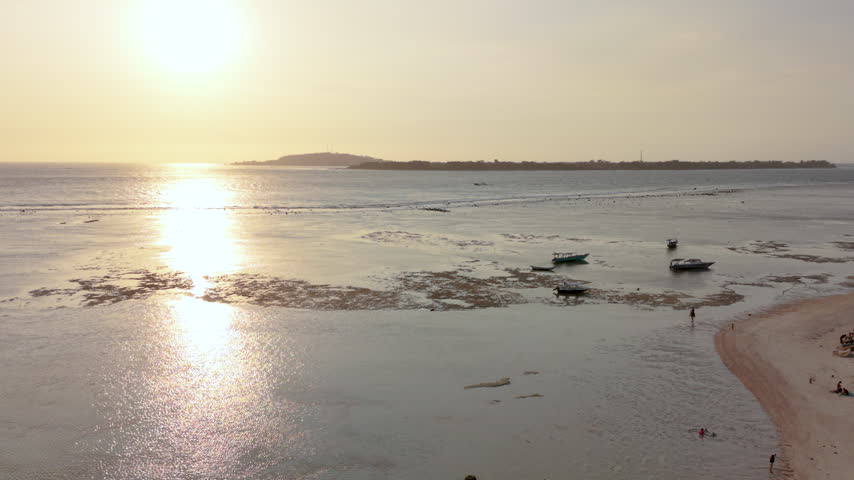 Aerial over shallow tidal waters off Gili Air, Indonesia at sunset, with sunlight reflecting across the calm sea, boats resting on the sandy shore at low tide, a horse rider on the beach and Gili Trawangan visible on the distant horizon, high-angle drone shot flying forward.