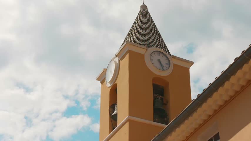 Low angle shot looking up at a yellow ochre church bell tower with clock face against a partly cloudy sky in the medieval village of Sainte-Agnès, French Riviera.