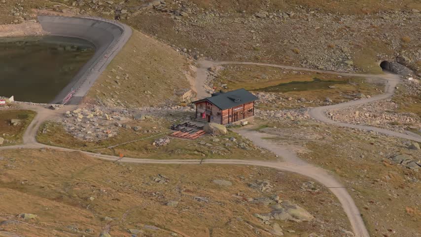 Aerial view of a mountain hut near the Aletsch Glacier with rocky terrain, a small reservoir, and winding dirt paths in Fieschertal, Valais, Switzerland.