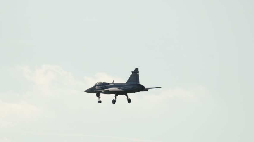 Military Saab JAS 39 Gripen fighter jet on final approach for landing. The aircraft lowers its landing gear while descending behind the trees