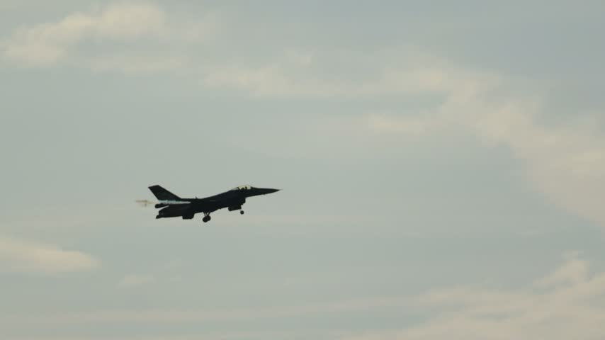 Silhouette of an F16 Fighting Falcon jet performing a barrel roll against a cloudy sky. The modern military aircraft showcases its agility