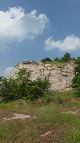 Vertical landscape view of rocky formations, trees and grass beneath a bright cloudy sky.