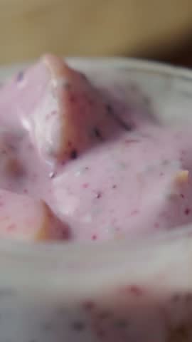 Creamy fruit yogurt with sweet berry bits is stirred with metal spoon inside glass cup, offering nutritious dessert texture while delicate dairy mixture drips slowly from kitchen utensil.