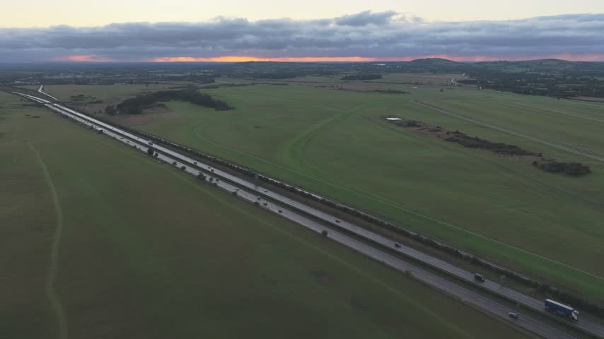 A long divided motorway cuts through open fields toward a distant tree line beneath a glowing evening horizon near Newbridge, County Kildare, Ireland.