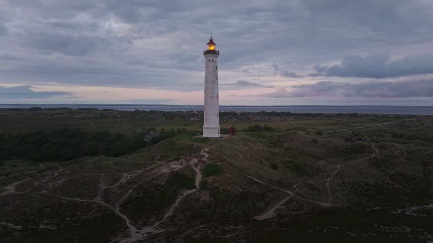Aerial view of the illuminated Lyngvig Lighthouse standing on a grassy dune under a cloudy twilight sky in Hvide Sande, Denmark.
