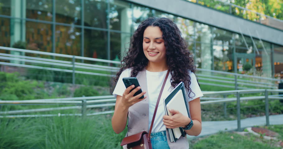 Portrait of happy smiling brunette girl student standing next to the building of the university while using mobile phone, outside