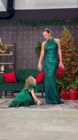 Woman helps a little girl stand beside a decorated Christmas tree during a festive holiday celebration.