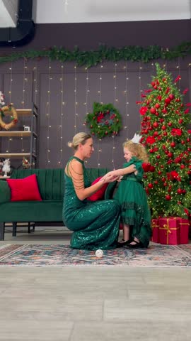 Woman helps little girl beside a decorated Christmas tree. Festive family interaction at home celebrates holiday tradition and childhood.