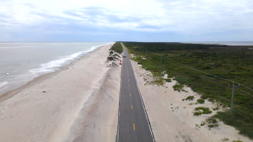 car approaches along a coastal road between dunes and Outer Banks vegetation.

