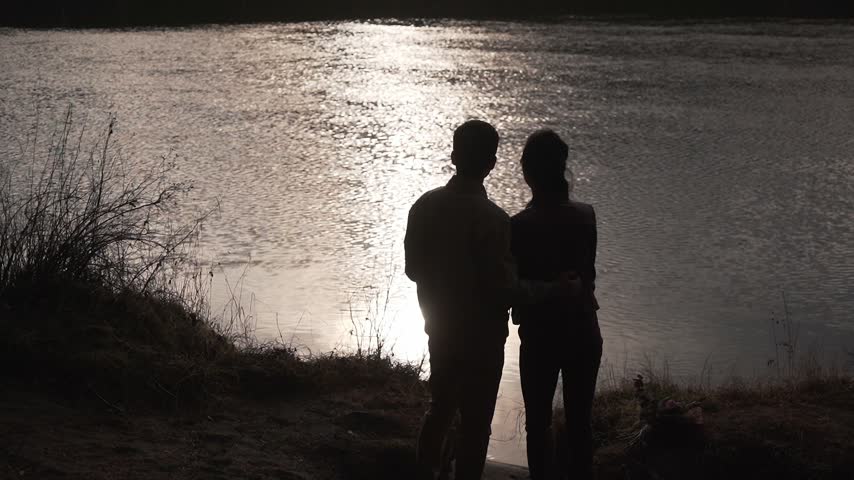 Dark silhouettes of a man and a woman embracing, standing on the riverbank with their backs to the camera at dusk after sunset. Love, intimacy, trust, expectation.