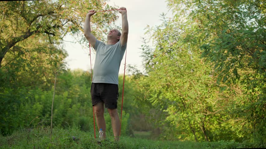 Adult athlete uses red resistance bands while exercising on a wooded outdoor trail, raising both hands during an upward pulling movement amid dense green vegetation and warm natural light