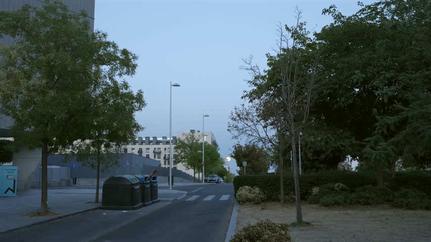 A serene urban street in Madrid, Spain, featuring trees, recycling bins, and a pedestrian crossing under a clear evening sky.