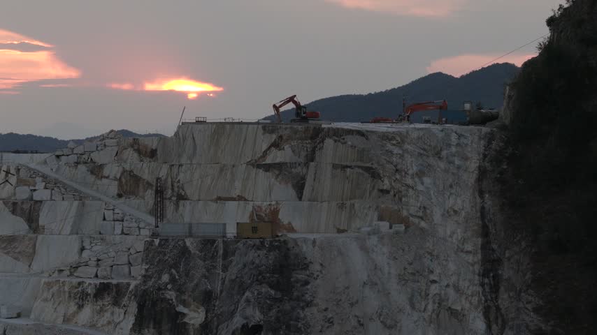 Aerial view of heavy machinery working in a white marble quarry during golden hour in Carrara, Tuscany, Italy.
