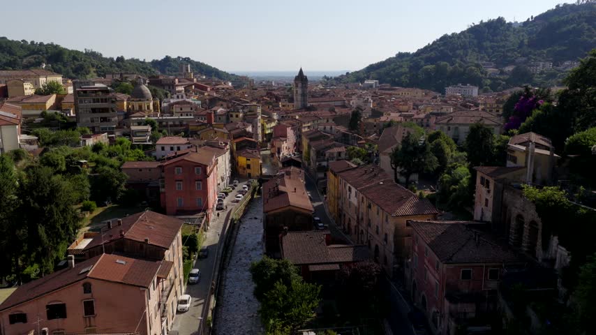 Aerial view of the historic city of Carrara with its traditional buildings and a river flowing through the center, set against lush hills in Carrara, Tuscany, Italy.