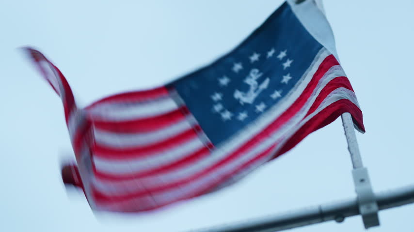 Close view of a United States yacht ensign in the wind. Red and white stripes ripple below a blue canton with white stars and an anchor above metal railings.