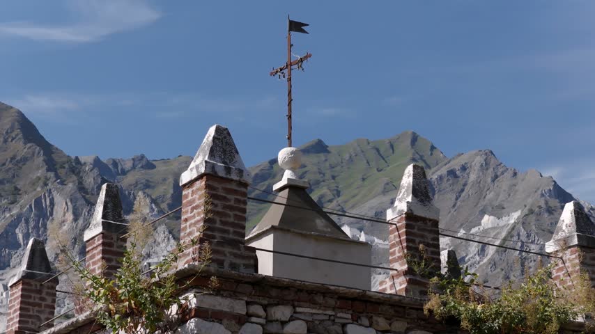 Aerial view of a stone structure with a decorative cross and weathervane set against the rugged Apuan Alps in Colonnata, Tuscany, Italy.