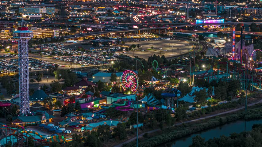 Aerial view of the illuminated Elitch Gardens amusement park with its glowing Ferris wheel and roller coasters set against city lights at night in Denver, Colorado, United States.
